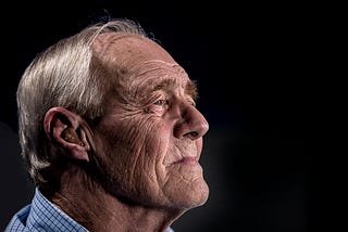 An old man with white hair, wrinkly face, and chequered shirt looking to the right