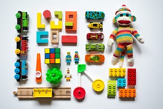 A set of colorful toys, including train cars, blocks, and hot wheels cars, is carefully arranged linearly in vertical and horizontal lines. There is also a sock monkey in a rainbow outfit.