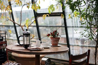 A small round table with 2 chairs by a window. On the table is a French press, coffee cups, and a potted plant. There are trees arching overhead.