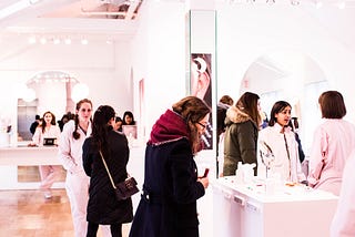 Customers browse a high-end looking cosmetics store with bright white lights as sales associates approach them to help with their shopping needs.