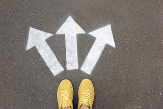 A pair of feet in yellow canvas sneakers standing in front of 3 arrows painted on the group that point in different directions.