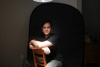 A man with dark hair & pale skin sits backwards on a chair on a dark room, lit dramatically from above.