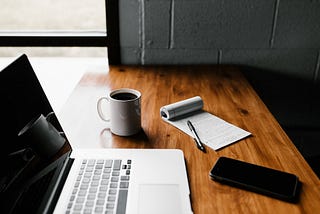 A laptop, coffee mug, pen, paper and mobile phone sitting on a table.