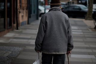 An old man walking with a stick, carrying shopping.