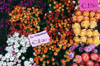 Image shows bunches of predominantly red, orange and yellow flowers in a flower market with pink pricing signs. The roses are 1.50Euro and the Rannunculas are 2Euros.