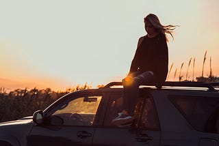 A blonde girl looks down as she sits on top of a black van in a field with a sunset in the background.