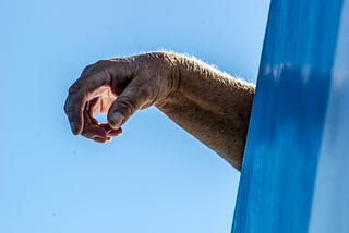 aging hand dangling over a blue wall against a clear blue sky