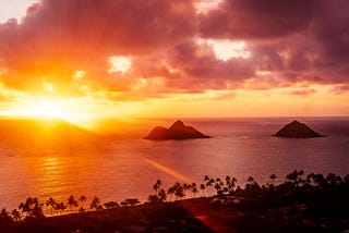 Photo of the ocean from a high vantage point. Two islands are visible, with deep orange and yellow colors of the sunrise.