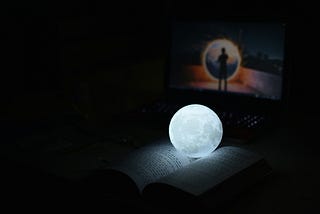 A glowing moon lamp laying on an open book, laptop in background