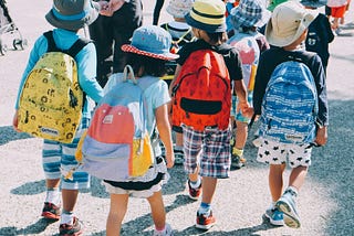 Young children with colorful backpacks pictured from the back as they walk away in pairs.