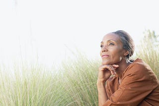 A Black woman, sitting in a grassy field and looking pensive.