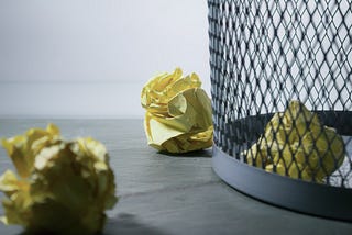 A photo showing a floor with three wrinkled yellow paper. There is a trashcan on the right, with one of the papers inside.