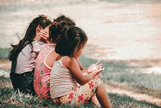 Three small children sit side by side on grass. The one closest to the camera is looking at her upturned hands