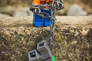 A lego figuring dangling from a cliff while another is helping him up