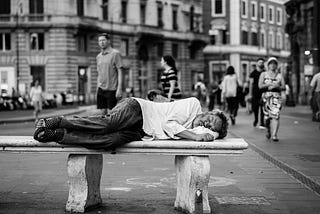 Homeless man sleeping on a stone bench while people walk around in the background.