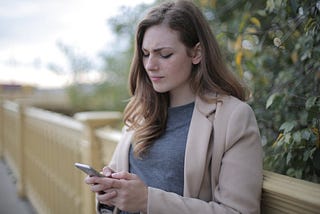 A white woman is standing on a bridge, looking at her phone and frowning.