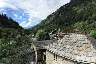 Image showing the brown tiled roof of a home in the foreground, with green trees and hillsides sloping downwards. Clouds hang ahead in the sky, with the Himalayan mountains slightly visible in the distance.