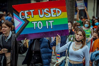 A protestor with the modern LGBTQIA+ Flag Sign that says “Get Used To It”