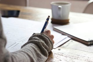 Someone’s arm holding a blue pen above pieces of paper, on a wooden surface. In the background, a notebook and a mug