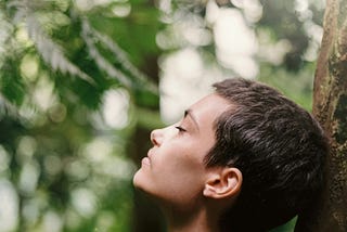 Lady with very short hair with eyes closed. Her head is slightly facing up and leaning against a tree.