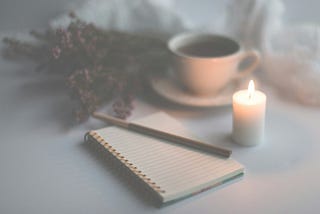 A notepad and pen next to a lit candle and cup of coffee
