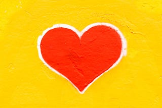 Red heart, with a white outline, painted on a bright yellow, stucco, wall