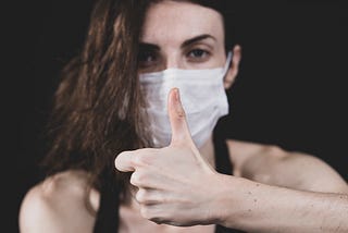A woman in a blue disposable face mask makes a thumbs-up gesture.