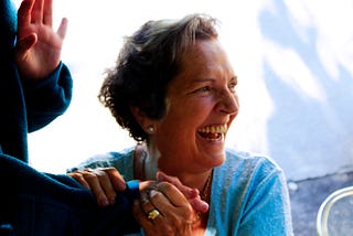 Older woman in blue cardigan, gripping someone’s hand and laughing.