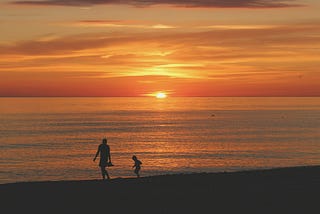 Just above the horizon a blazing orange sky hovers above a beach, where a man and a boy walk, the boy lagging or following as he would.