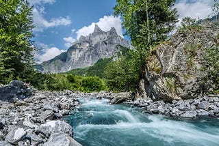 A fast-flowing river with trees and a mountain in the background, against a blue sky dotted with clouds.