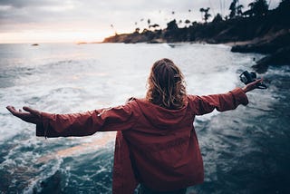 White person with medium-length brown hair in a red hooded jacket, arms outstretched in openness as they look out into the cove of an ocean side.