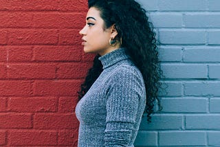 A woman with curly dark hair stands in profile against a wall split into red and blue sections, wearing a grey long-sleeve top and black pants.
