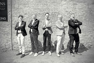 Black and white photo of five men in sport jackets with their arms crossed.