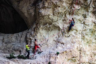 A person climbing a rock face on the upper right with two people holding ropes on the lower left