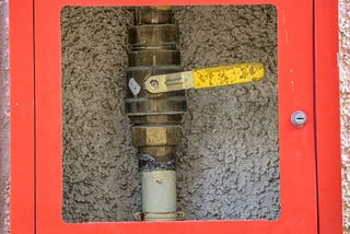 Photo of metallic gas pipe with a valve, in a red security box, the box center has a glass and therefore is transparent, so the valve is visible,