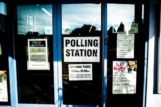 Entrance of a polling station during the day with some informative flyers stuck from inside of the facility, this includes a paper saying ‘POLLING STATION’.