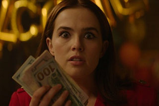 Zooey Deutch as Peg stares into the camera with wide eyes as she holds a stack of $100 bills.