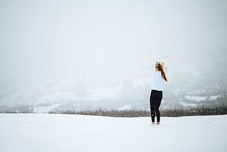 Young woman in black pants and white jacket standing at the precipice of a white glacier.