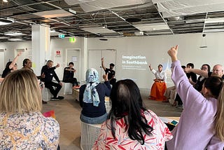 A group of people sit in a circle on stools with their thumbs up in the air, conveying that they think something is true.