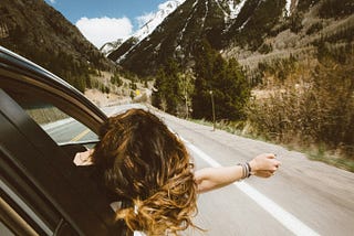 girl sticking her head and arm out the window in a car on a road trip driving through a scenic view of the mountains