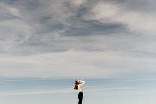 A woman stands on sand with her palms covering her face and her head raised to the sky