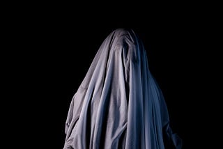 The top half of a figure, probably a woman’s, with its back to the camera, shrouded by a veil or cloth, against a black background