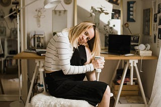 A woman, middle-aged, sitting by her desk, coffee in hand. There is a laptop with a black screen in front of her and the background of her room feels very ethereal.