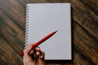 Photo of a white hand holding a red pen over a blank notebook set on a wooden background