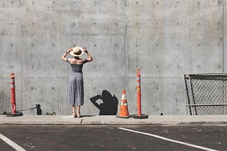 Woman in dress and straw hat with back to camera stares at tall concrete wall. Orange traffic cones and fencing nearby.