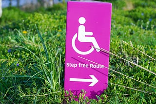 A pink sign on some grass, with a wheelchair symbol, the words “step free route” and an arrow to the right.