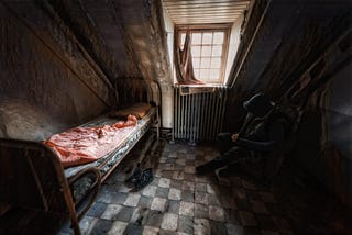 A dark destroyed room with a filthy bed, grimey walls, and a dirty floor. A lone figure in a black hoodie sits in a chair in the corner of the room looking at the floor with his shoulders slouched.