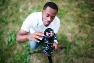 A person crouches outdoors on grass, adjusting a camera mounted on a tripod. The focus is on the camera lens in the foreground, with the photographer slightly blurred in the background.