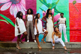 6 Black women dressed in varying shades of white posed against a brick wall painted in colorful flowers