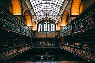 A library filled with bookcases of books on either side. A glass ceiling, criss-crossed with frames lets in a little light.